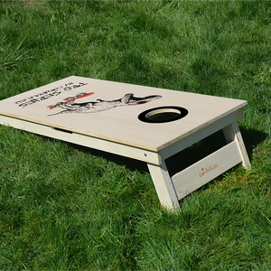 cornhole Pro de compétition t-rex sur son skateboard, sur le gazon, vue de derrière