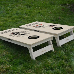 cornhole de compétition deux planches t-rex sur son skate et tête de mort, sur le gazon, vue de derrière